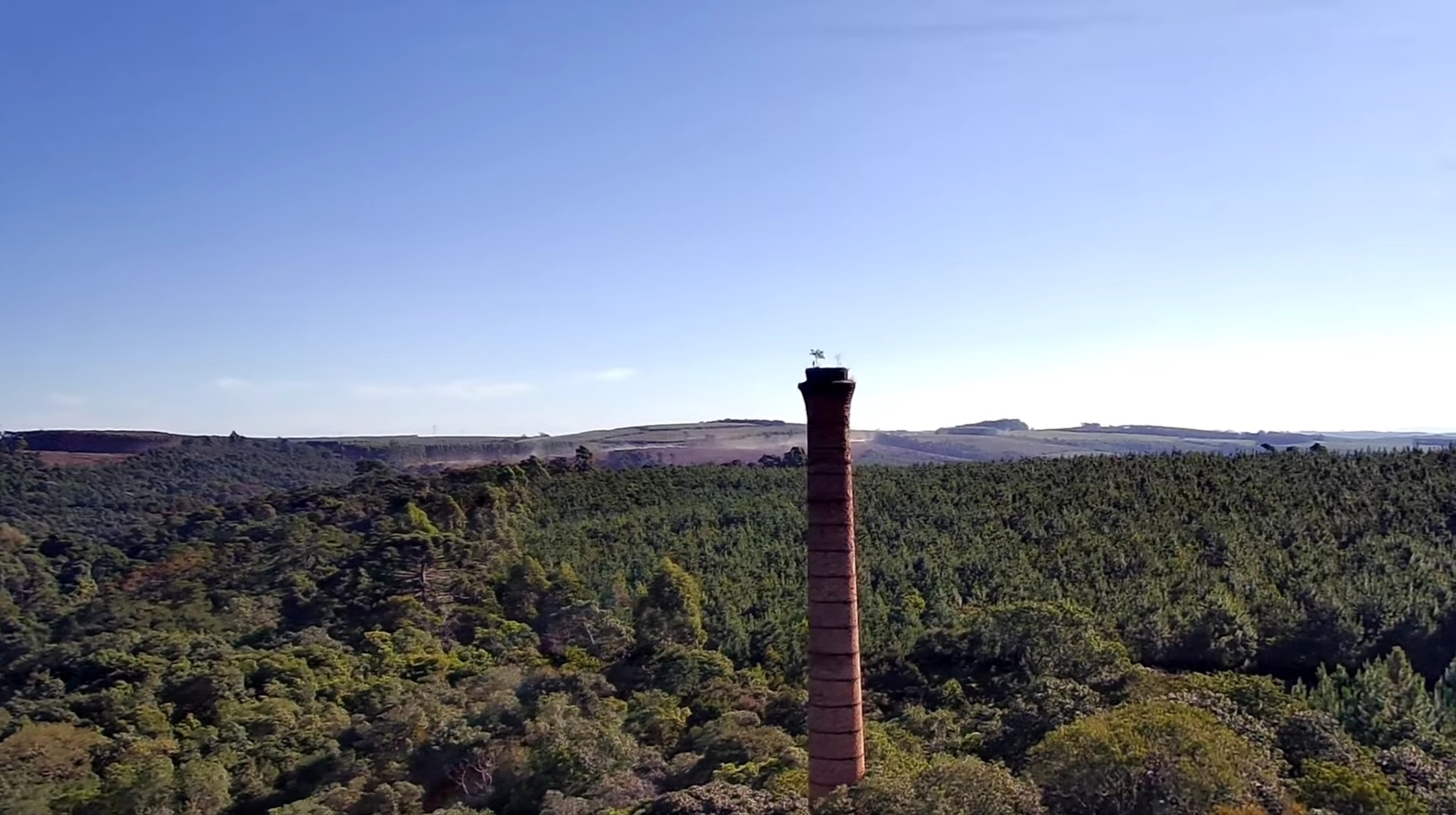 Vista distante da chaminé da antiga fábrica da Klabin em Harmonia, Telêmaco Borba, mostrando a integração da estrutura industrial com a natureza.