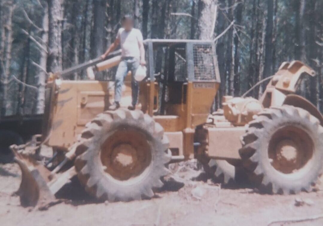 Fotografia antiga de época mostrando um grande trator florestal articulado do tipo skidder, de cor amarela, estacionado em uma área de terra batida. Um operador com o rosto desfocado está em pé sobre o grande pneu dianteiro esquerdo da máquina. Ao fundo, há uma floresta densa com troncos finos e altos de árvores de pinus sob a luz do dia.
