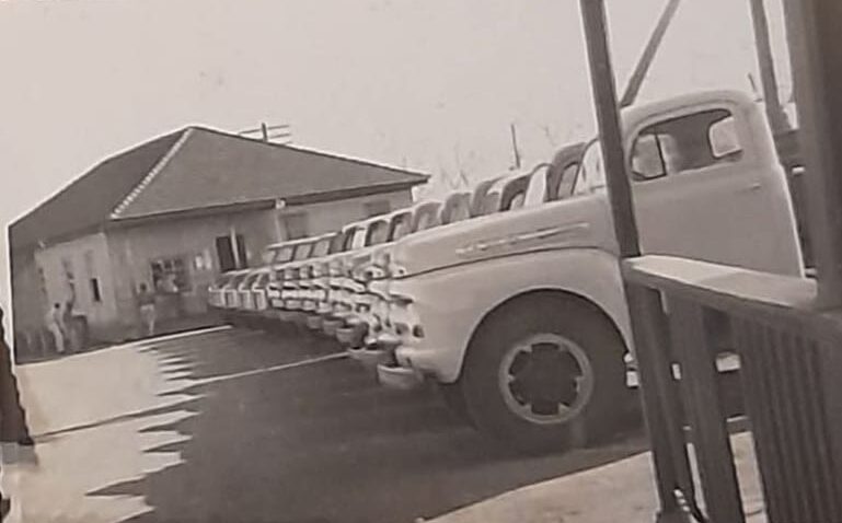 Fotografia antiga em tons de sépia mostrando uma grande frota de caminhões Ford clássicos dos anos 1950 alinhados em um pátio de paragem. Em primeiro plano, destaca-se a lateral branca dos veículos estacionados em diagonal. Ao fundo, há uma construção de alvenaria antiga com telhado escuro, registrando a infraestrutura de transporte dos antigos acampamentos.