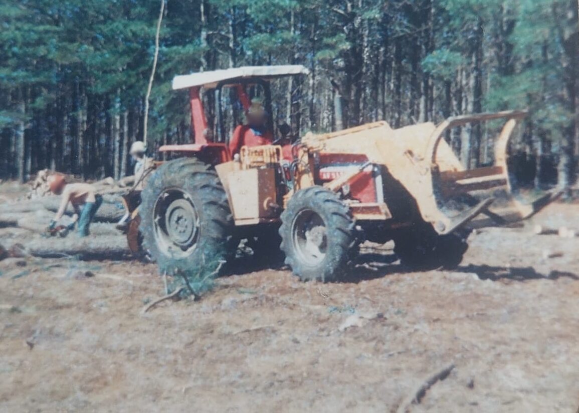 Fotografia antiga de época mostrando um trator florestal de cor vermelha com capota branca, equipado com uma garra carregadeira amarela na parte frontal. O trator está estacionado em um terreno de terra batida com restos de galhos. Ao fundo, no canto esquerdo, um trabalhador opera uma motosserra sobre troncos de madeira, com uma floresta densa de pinus completando o cenário.