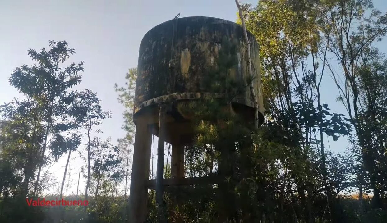 Fotografia colorida mostrando uma antiga caixa d'água cilíndrica de concreto suspensa por pilares altos. A estrutura apresenta marcas severas de desgaste e umidade pelo tempo, estando cercada por árvores altas e vegetação nativa sob um céu claro, com o nome Valdecirbueno escrito em vermelho no canto inferior esquerdo.
