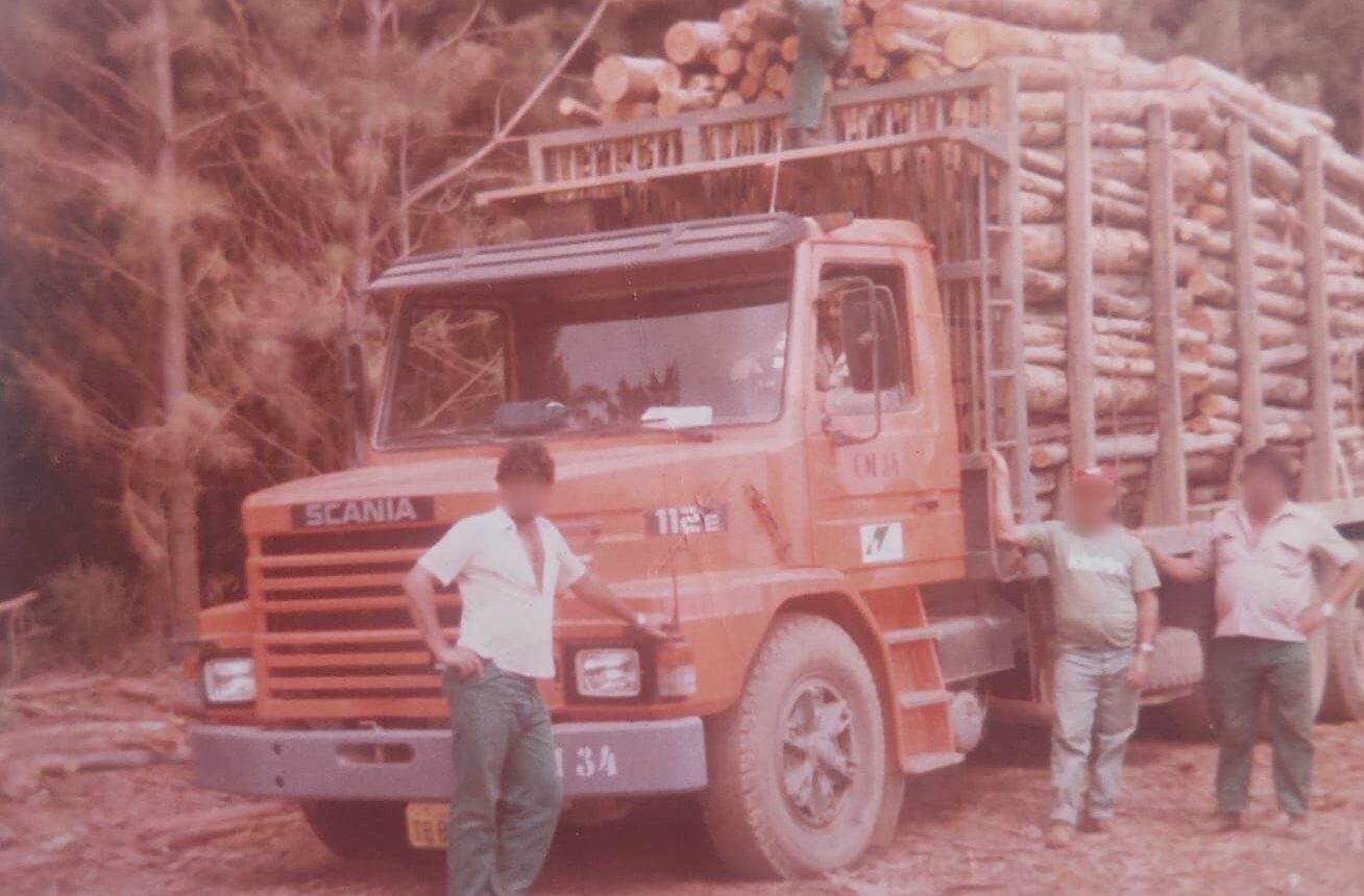 Fotografia antiga de época mostrando um caminhão Scania T112 bicudo de cor laranja, carregado com toras de madeira reflorestada em uma área de corte. Três trabalhadores posam em frente e ao lado do veículo, enquanto um quarto homem aparece sobre a carga de madeira. Ao fundo, destacam-se árvores de pinus em um ambiente florestal rústico.