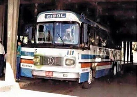 Fotografia antiga de um ônibus clássico da marca Mercedes-Benz com carroceria Ciferal, pintado nas cores branca, azul e faixas laranjas, estacionado em uma plataforma de embarque coberta. Há passageiros visíveis nas janelas e pessoas na plataforma ao lado do veículo, representando o transporte dos trabalhadores e atletas da região.
