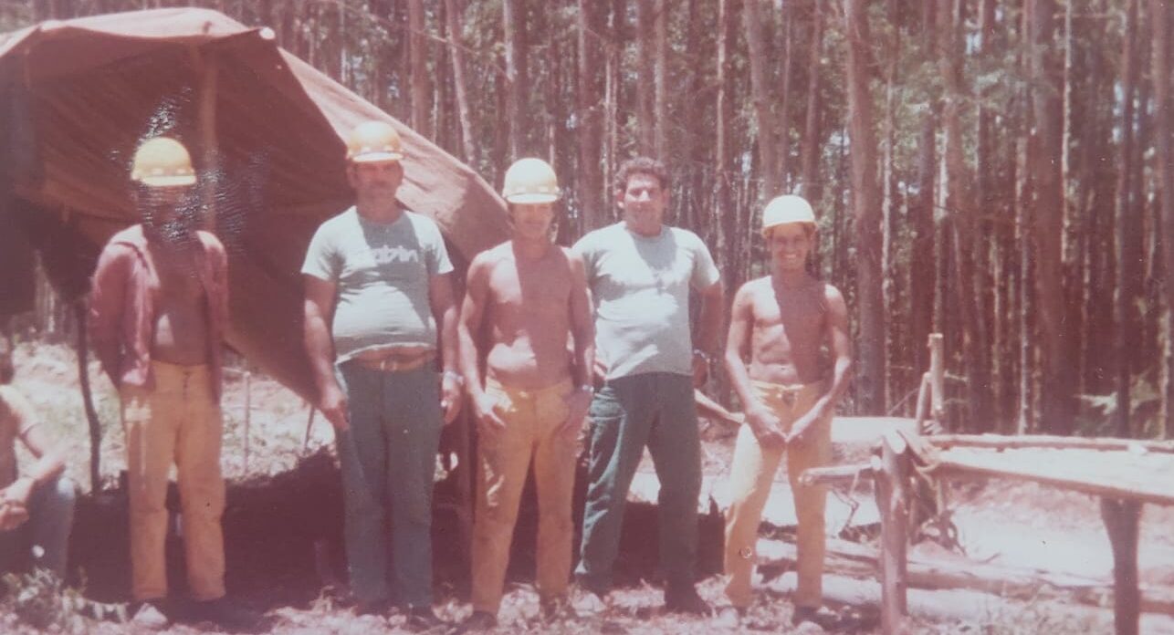 Fotografia antiga em tom envelhecido mostrando cinco trabalhadores florestais posando em pé em meio a uma plantação de pinheiros. Eles usam capacetes de proteção amarelos e brancos, e um deles veste uma camiseta com o logotipo antigo da Klabin. Ao fundo, há uma barraca de lona improvisada na mata, registrando o cotidiano dos operários do reflorestamento.