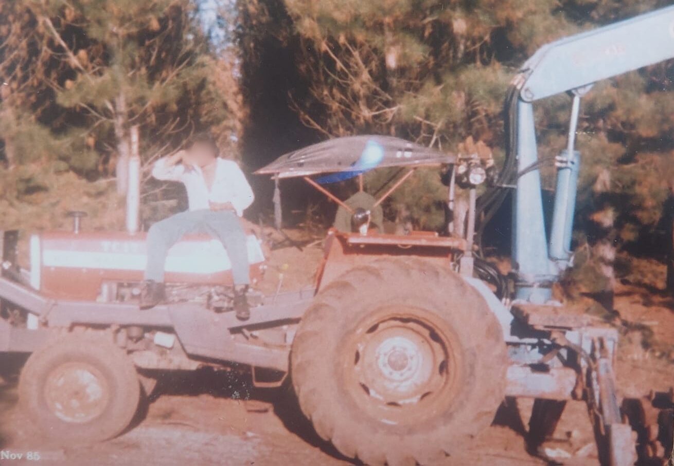 Fotografia antiga de época, datada de novembro de 1985, mostrando um trator florestal vermelho equipado com um braço hidráulico azul na parte traseira. Um jovem trabalhador, com o rosto desfocado, está sentado sobre o capô do veículo, que possui uma pequena cobertura rústica sobre o assento do operador. Ao fundo, há uma área com árvores de pinus sob a luz do sol.