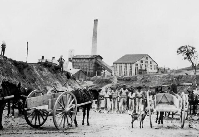 Fotografia histórica em preto e branco mostrando operários e carroças de tração animal em um canteiro de obras de terra batida. Em primeiro plano, destacam-se duas carroças de madeira com grandes rodas de raios atreladas a cavalos, além de um grupo de trabalhadores alinhados ao centro e um pequeno bode no chão. Ao fundo, avista-se o complexo de uma fábrica antiga com grandes galpões e uma chaminé industrial alta sob um céu claro.