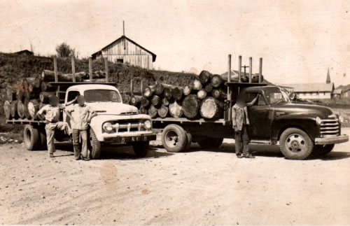 Fotografia histórica em tom sépia mostrando dois caminhões antigos dos anos 1950 carregados com toras de madeira em um terreno de terra batida. À esquerda, um caminhão Ford de cabine clara com dois homens em pé ao lado; à direita, um caminhão Chevrolet de cabine escura com um motorista apoiado na porta. Ao fundo, avistam-se casas de madeira em uma colina e a torre de uma igreja sob um céu claro.