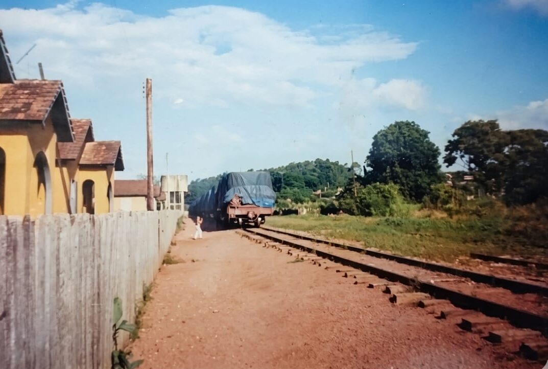 Fotografia antiga de uma linha de trem de ferro que corta um acampamento ou vila operária. À esquerda, há uma sequência de casas amarelas históricas com fachadas idênticas atrás de uma cerca de madeira. No centro, vagões de carga cobertos por uma lona azul estão sobre os trilhos, com uma estrada de terra ao lado e vegetação verde ao fundo sob um céu com nuvens.
