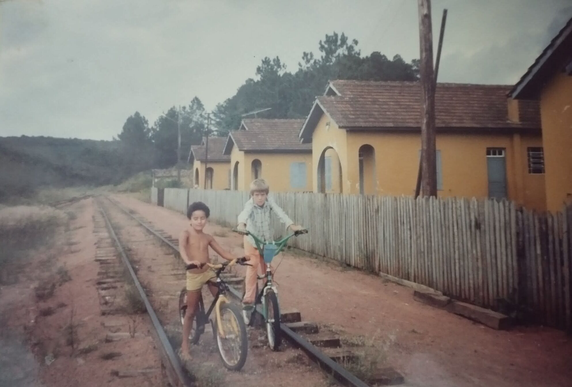 Fotografia antiga mostrando dois meninos andando de bicicleta sobre os trilhos de uma linha de trem desativada em uma vila operária. Ao fundo, vê-se a sequência de casas amarelas antigas com cercas de madeira e uma área de floresta com pinheiros sob um céu nublado, registrando a infância nos antigos acampamentos.