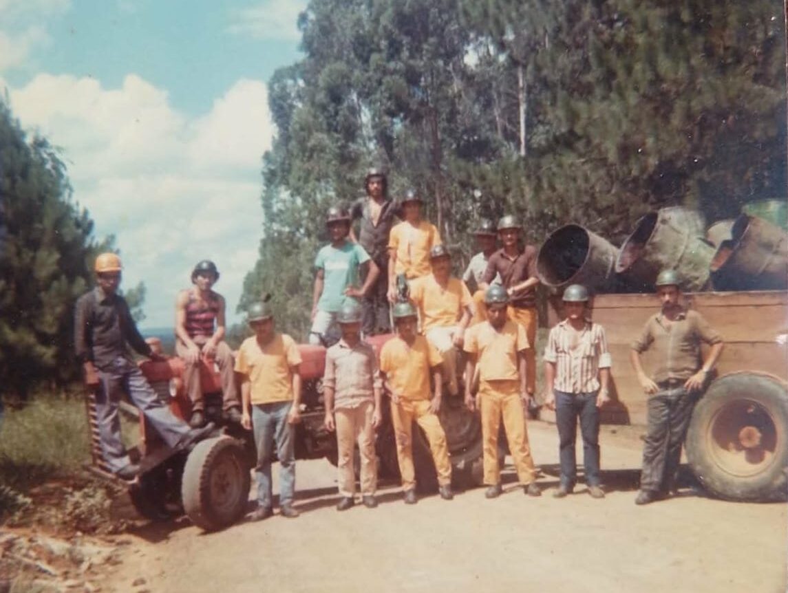 Fotografia antiga mostrando um grupo de operários florestais posando para foto ao redor de um trator vermelho em uma estrada de terra. Vários homens usam capacetes de proteção, alguns estão sentados no trator e outros em pé ao lado da carretinha de madeira carregada com grandes tambores de metal, com uma floresta de pinheiros ao fundo.