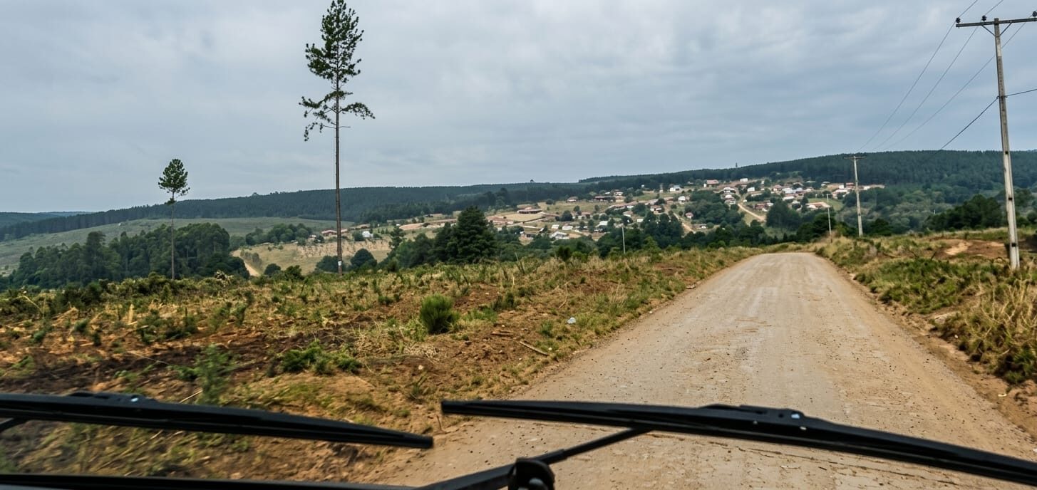 Fotografia colorida tirada de dentro de um veículo, mostrando a perspectiva de uma estrada de terra que leva a uma vila com várias casas no topo de uma colina rústica. Ao redor do caminho, há vegetação rasteira, árvores de pinus altas e postes de energia elétrica, com uma vasta floresta de reflorestamento sob um céu nublado ao fundo.