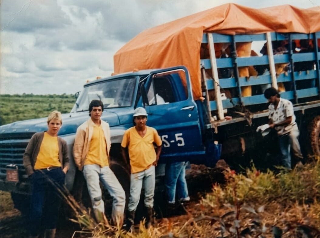 Fotografia antiga mostrando um caminhão Ford azul com carroceria de madeira e lona laranja na caçamba, usado para o transporte de trabalhadores. Três rapazes posam em pé apoiados na frente do veículo em uma estrada de terra, enquanto outros homens aparecem na caçamba e na lateral do caminhão, registrando o cotidiano dos operários rurais.