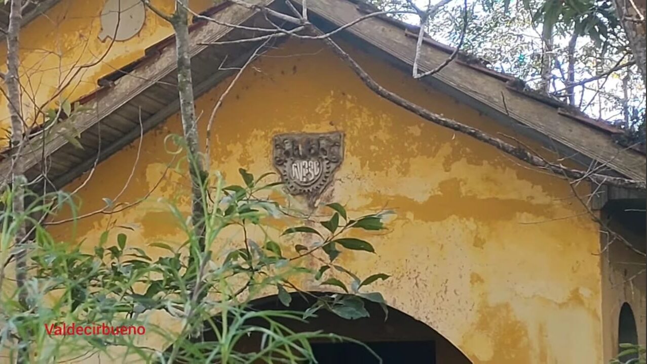 Fotografia colorida em plano fechado mostrando o topo da fachada de uma antiga casa amarela abandonada de uma vila ferroviária. A pintura está desgastada e descascada pelo tempo, com galhos e plantas cobrindo parte da estrutura. No centro, acima de um arco de entrada, destaca-se um brasão em relevo com a sigla RVPSC, com o nome Valdecirbueno em vermelho no canto inferior esquerdo.