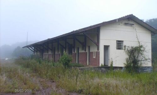 Fotografia colorida mostrando uma antiga estação ferroviária ou armazém abandonado em um dia de forte neblina. O prédio comprido possui paredes brancas e uma base de tijolos vermelhos, com um telhado estendido sustentado por vigas de madeira inclinadas. Há mato alto e vegetação rasteira crescendo ao redor da estrutura, com a data 05/02/2008 marcada em cinza no canto inferior esquerdo.