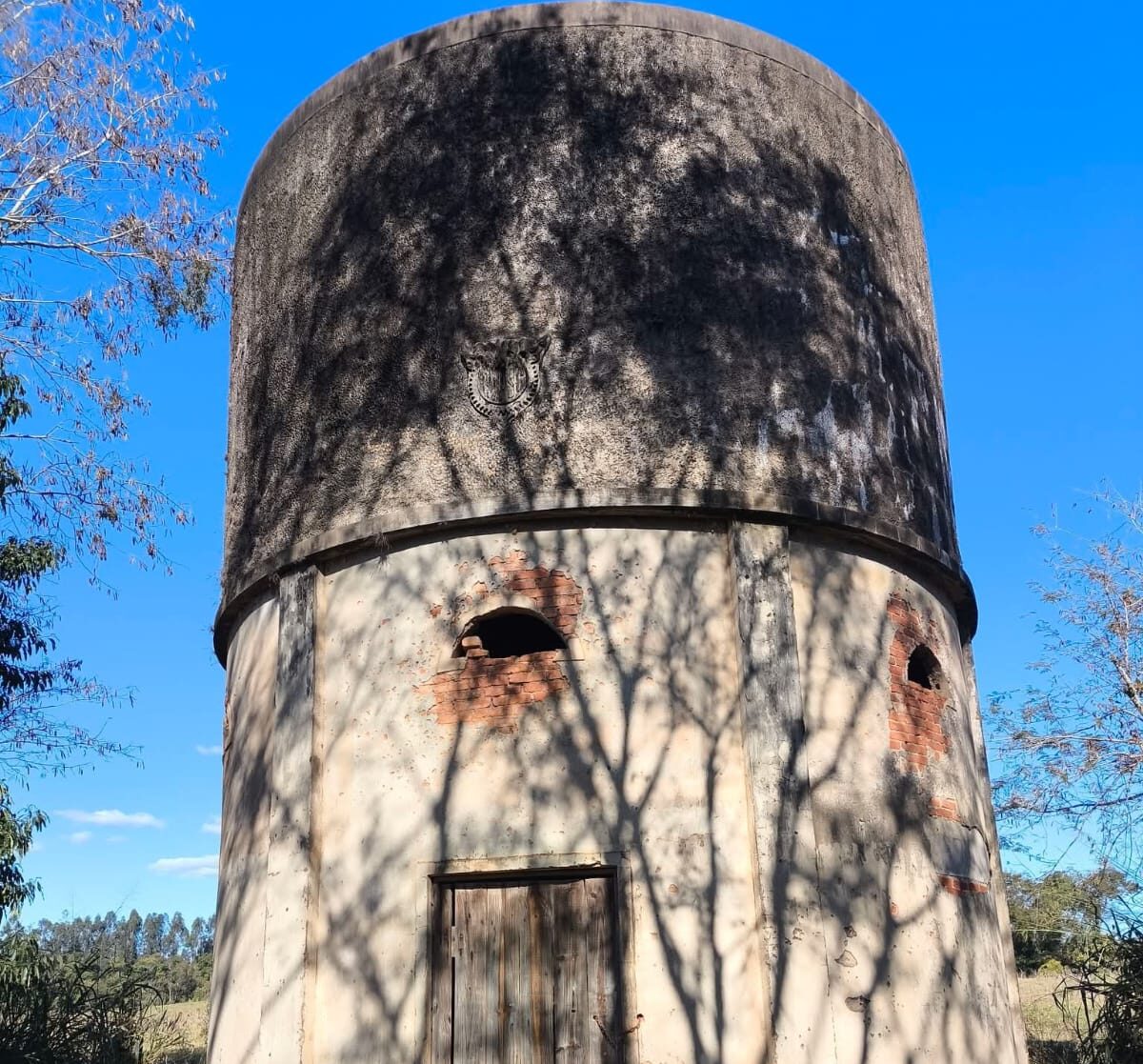 Fotografia colorida mostrando uma antiga torre ou caixa d'água cilíndrica de concreto da ferrovia, sob um céu azul. A estrutura possui marcas do tempo, uma porta de madeira fechada na base e pequenas aberturas em arco com tijolos vermelhos expostos. Na parte superior de concreto escuro, destaca-se o brasão em relevo da RVPSC, com sombras de galhos de árvores projetadas em toda a fachada.