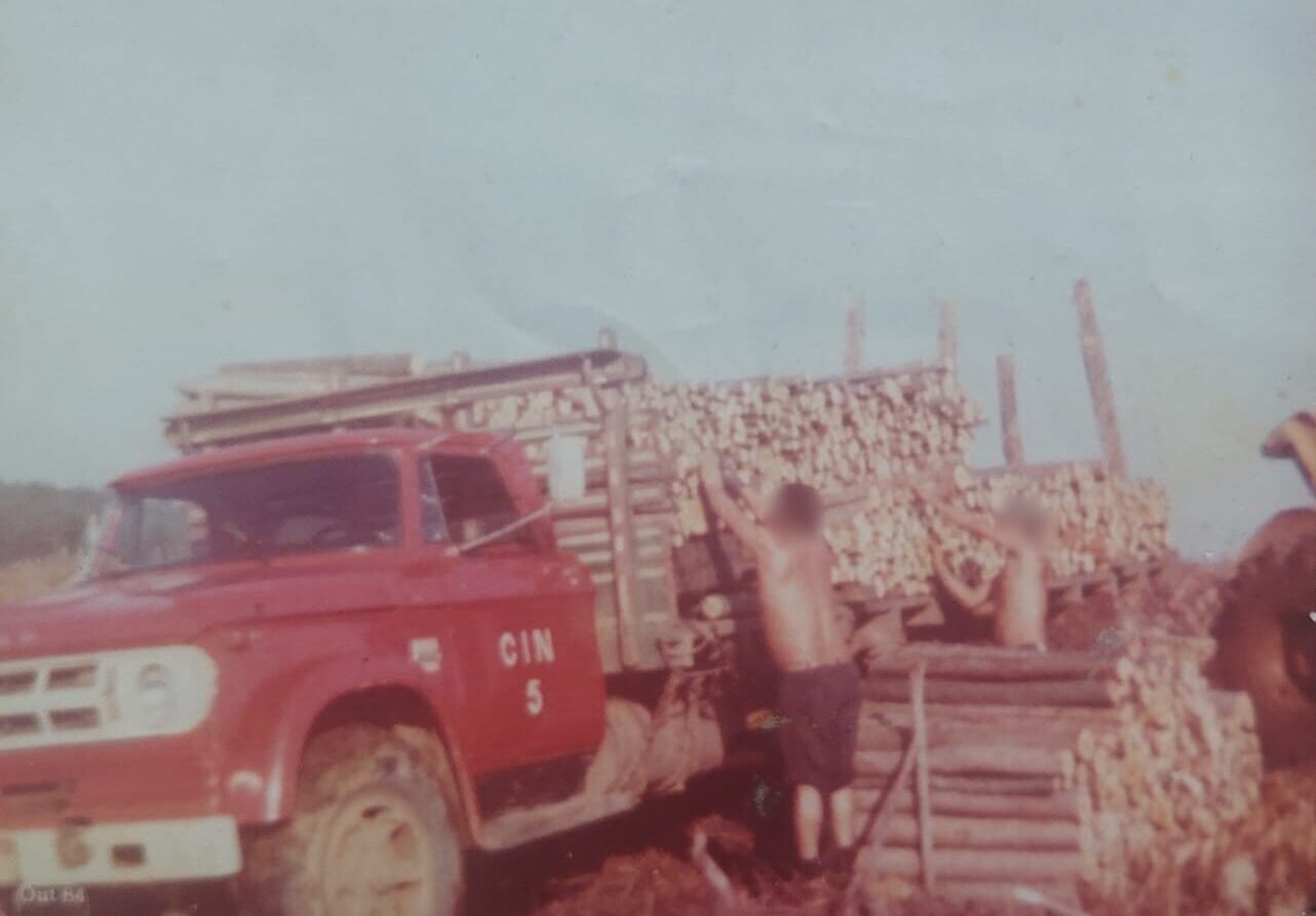 Fotografia antiga de época, datada de outubro de 1984, mostrando um caminhão antigo vermelho Chevrolet D60 sendo carregado manualmente com toras de madeira rústica. Dois trabalhadores sem camisa aparecem empilhando os troncos na carroceria do veículo. No chão, há outra grande pilha de madeira cortada sob a luz do dia.