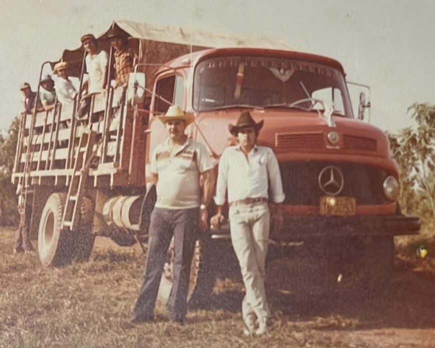 Fotografia antiga de época mostrando um caminhão Mercedes-Benz de cor vermelha com carroceria de madeira adaptada para o transporte de trabalhadores rurais. Vários homens aparecem acomodados na carroceria sob uma cobertura de lona, enquanto dois homens vestindo chapéus posam em pé em frente à cabine do veículo. O caminhão está estacionado em um terreno de vegetação rasteira sob a luz do dia.