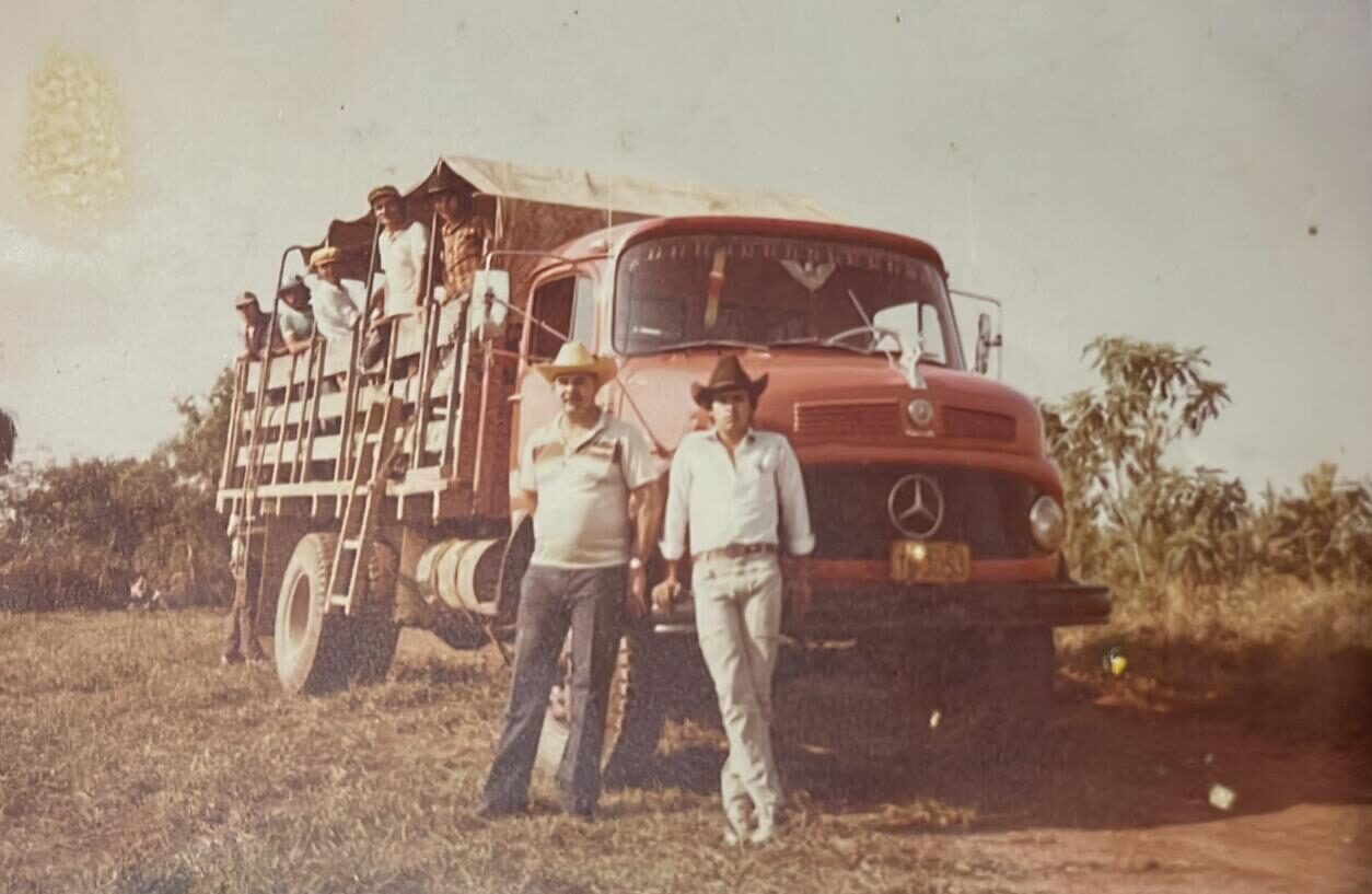 Fotografia antiga em tom envelhecido mostrando trabalhadores rurais em um caminhão Mercedes-Benz vermelho com carroceria de madeira. Vários homens estão em pé na caçamba do veículo e, em primeiro plano, dois homens usando chapéus de caubói posam em pé ao lado da cabine, registrando o transporte de operários nos acampamentos.