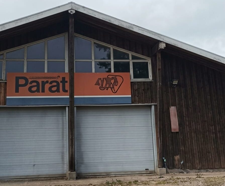 Fotografia colorida da fachada de um grande galpão rústico de madeira escura com duas grandes portas de metal cinzas fechadas. Acima das portas, há uma placa horizontal nas cores laranja e azul com o nome Parat escrito em preto e um logotipo mecânico estilizado ao lado. Na parte superior, há janelas de vidro sob a estrutura do telhado, com o céu nublado ao fundo.